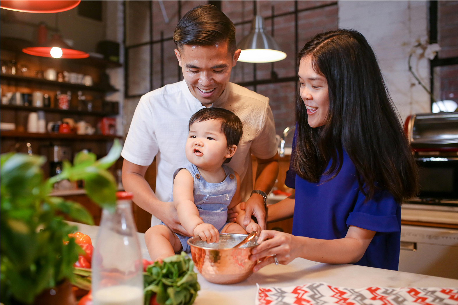 An adult couple and a baby are in a kitchen in Santa Clarita. The baby sits in a mixing bowl on the counter while the adults smile and interact with the child. Various kitchen items are visible around them.
