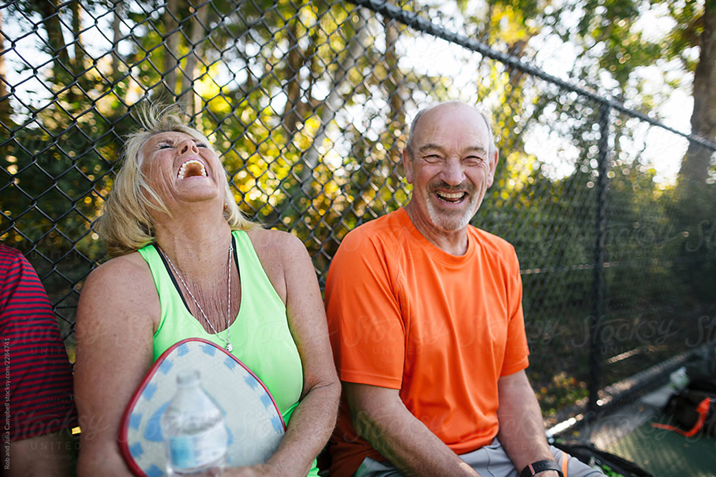 Two older adults sit on a bench by a chain-link fence in Santa Clarita, holding pickleball paddles and laughing together outdoors in the bright sunlight.