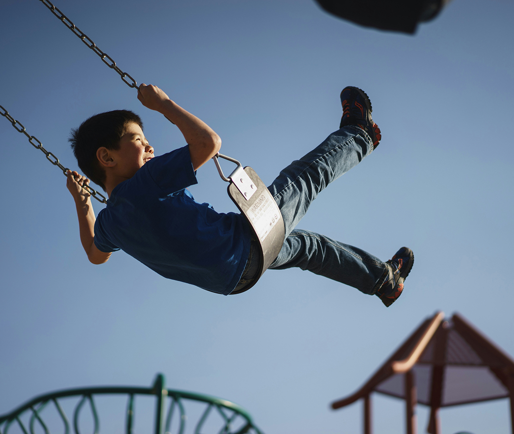 A young boy in a blue shirt and jeans swings high on a playground swing under the clear blue sky of Santa Clarita Valley.
