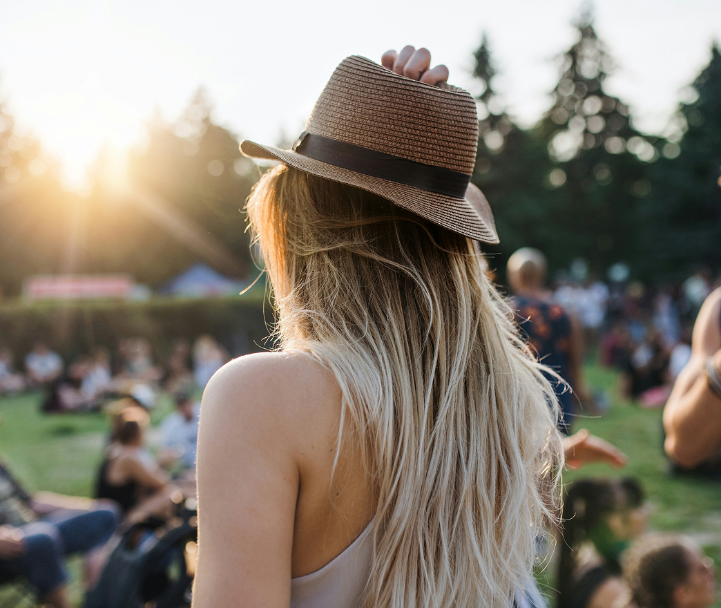 A person with long blonde hair and a brown hat stands outdoors at a sunlit Santa Clarita Valley event, surrounded by a crowd sitting on grass.