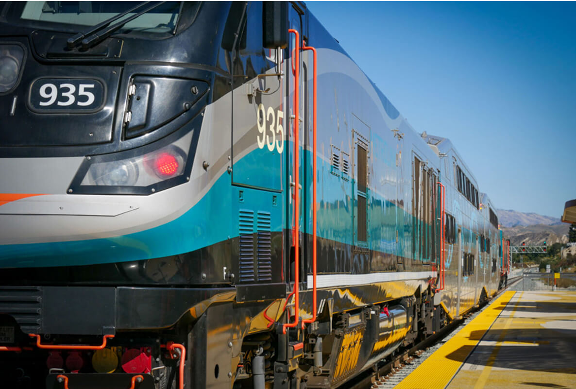 A modern passenger train numbered 935 is stopped at a yellow-lined platform under clear skies, with mountains of the Santa Clarita Valley visible in the background.