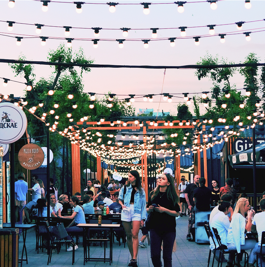 People dine and walk beneath string lights at an outdoor restaurant area in Santa Clarita Valley. Tables are filled, and the atmosphere is lively with greenery and decorations overhead.