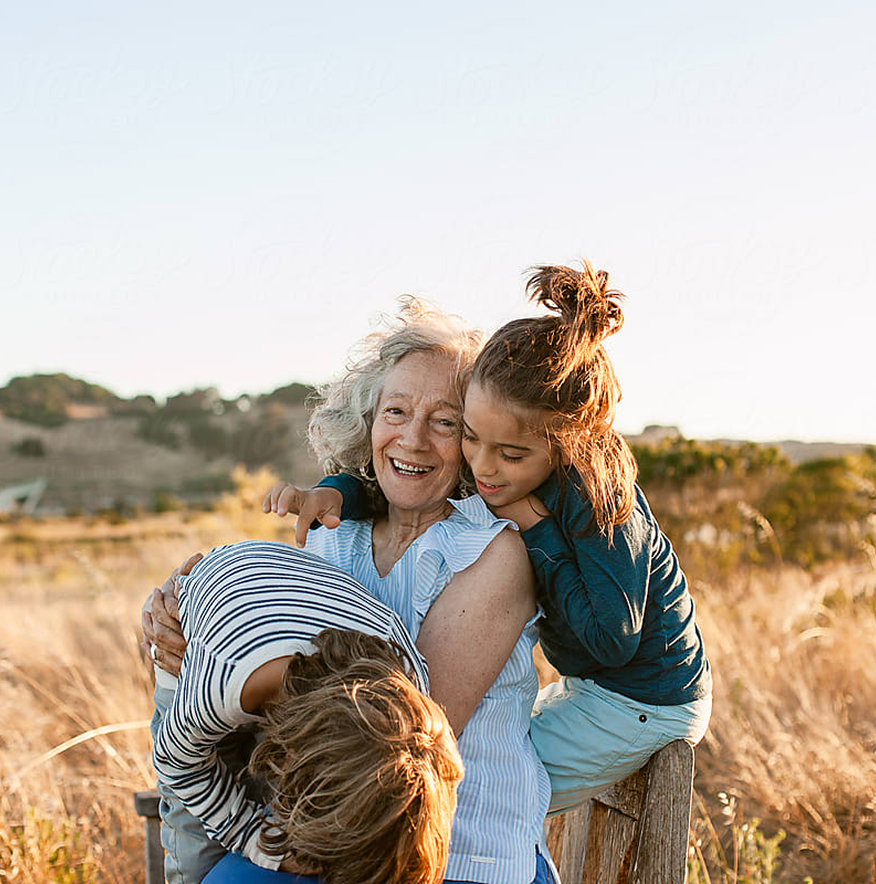 An older woman sits outdoors in a Santa Clarita Valley field, smiling as two children hug and lean on her, with sunlight illuminating the scene.