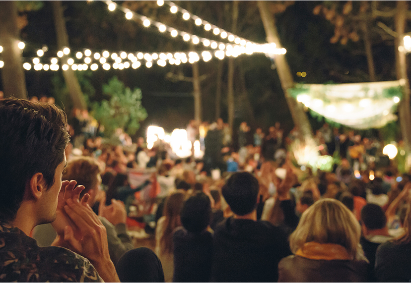 A crowd of people sits outdoors under string lights in Santa Clarita, facing a stage area, with trees and dim lighting creating a festive atmosphere.