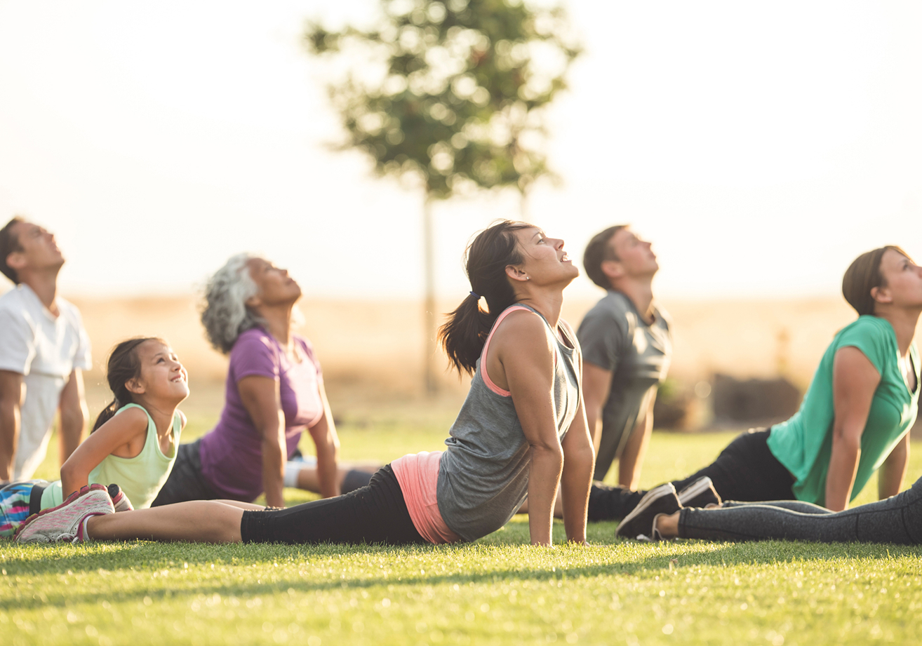A group of people of various ages practicing yoga outdoors on grass in Santa Clarita, performing the upward-facing dog pose, with a tree and open field in the background.